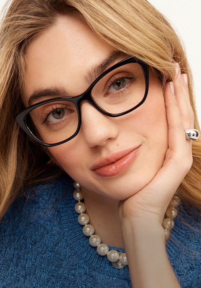 Woman wearing black glasses with a blue sweater and pearl necklace on a white background