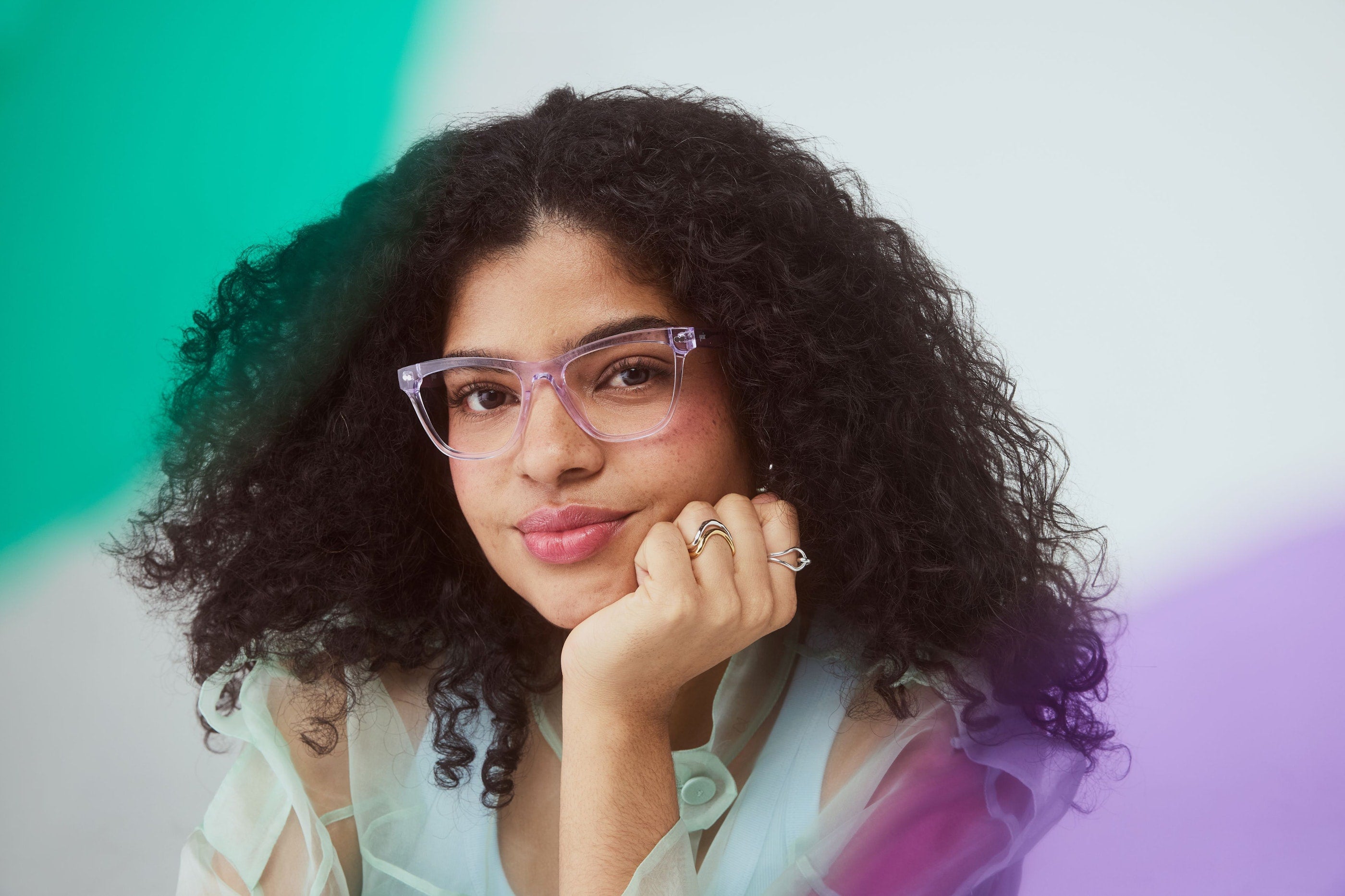 A woman wearing lilac Pair glasses, resting her chin on her hand against a green and purple backdrop